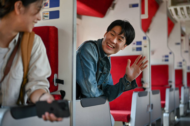 フレンドリーでハンサムな若いアジアの男性バックパッカーや旅行者は、電車の中で旅行中に新しい友人を迎えるために彼の手を振っています。友情と旅のコンセプト - 写真・画像