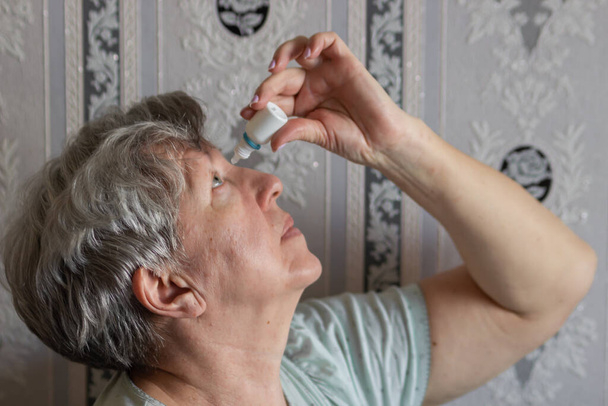 Senior woman dripping some medicine with dropper on her eyes at home. Old female using bottle of eyedrops. Sick lady suffering from irritated eye, optical symptoms. Eye health and treatment concept. - Photo, Image