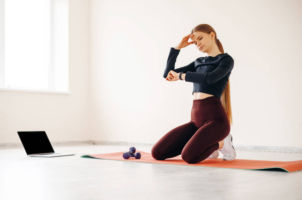 Tired woman having rest at yoga mat after workout. Tired and exhausted female athlete sitting on floor at gym with a two dumbbells and measures the pressure on the fitness bracelet - Photo, Image
