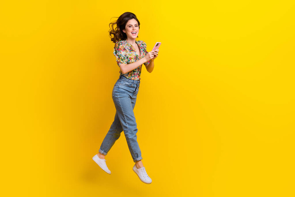 Full body cadre of young girl jumping wear flowers print blouse jump use phone internet connection fast speed isolated on yellow background. - 写真・画像