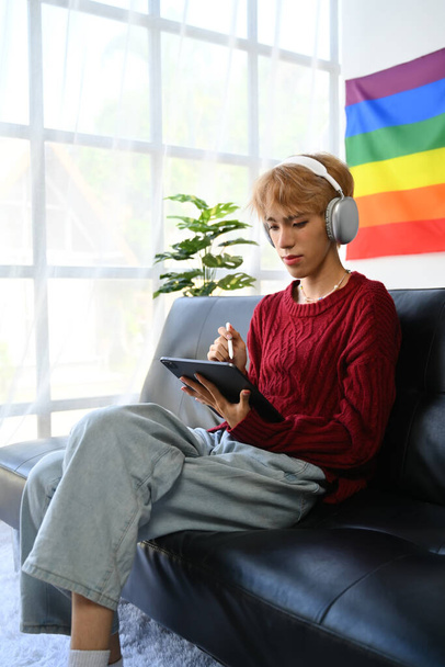 Portrait of casual teenage gay man wearing headphone using digital tablet on couch in living room with pride rainbow flag on wall. - Foto, imagen
