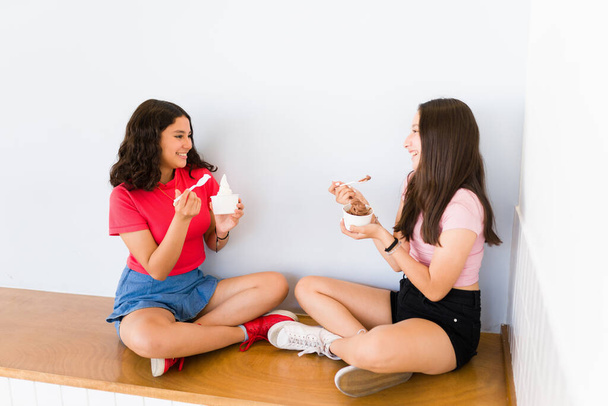 Adolescentes hispanas hablando de chismes y sonriendo felices mientras comen delicioso helado o yogur congelado  - Foto, imagen