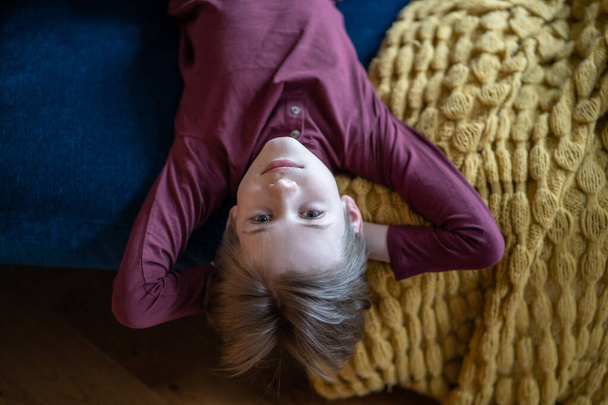 Portrait teenage boy lying on sofa upside down with hands behind head and looking camera. Smile on child face. Son resting before after school. International Children Day - Photo, Image