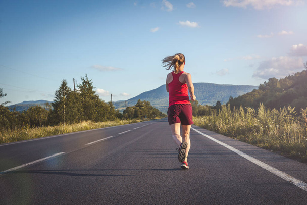 Fitness blonde woman running on the asphalt road at the mountains. Dramatic sky - Foto, afbeelding