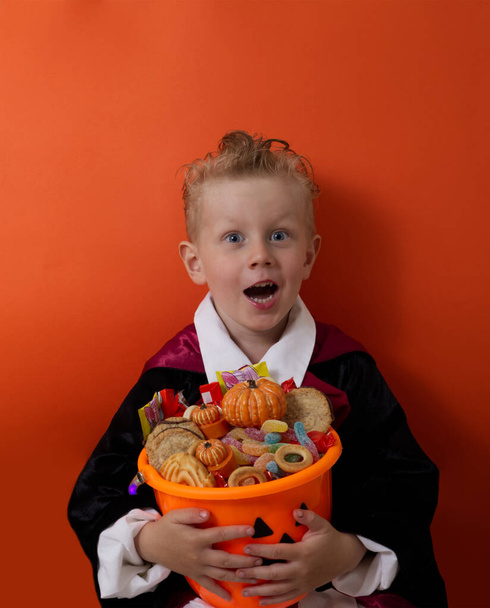 Ein Junge im Vampirkostüm hält einen Eimer mit Süßigkeiten auf orangefarbenem Hintergrund. - Foto, Bild