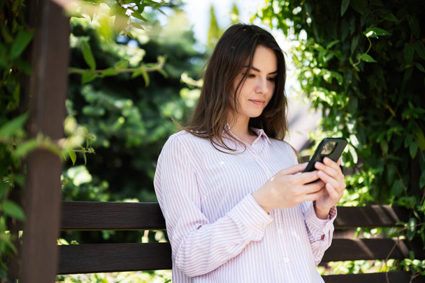 Молодая фрилансер женщина в рубашке сидеть на скамейке в летнем парке на открытом воздухе отдыха и использовать мобильный телефон.  - Фото, изображение