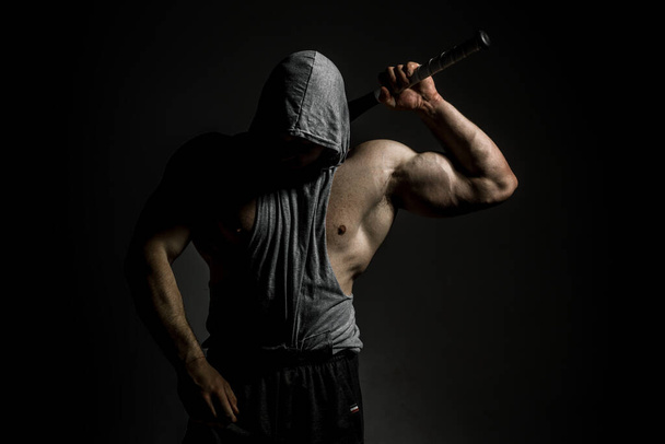 Dangerous muscular man in the mask with baseball bat ready for fight. An aggressive hooded man is holding a baseball bat on black - Foto, Imagem