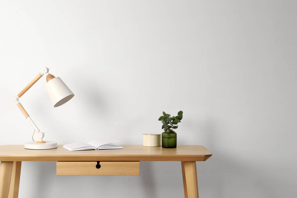 Stylish modern desk lamp, open book and plant on wooden table near white wall, space for text - Foto, afbeelding