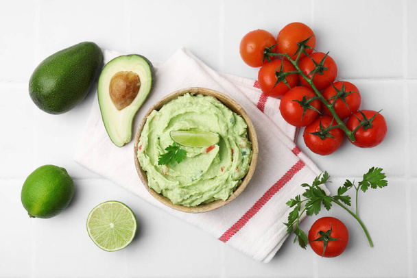 Bowl of delicious guacamole and ingredients on white tiled table, flat lay - Foto, Bild