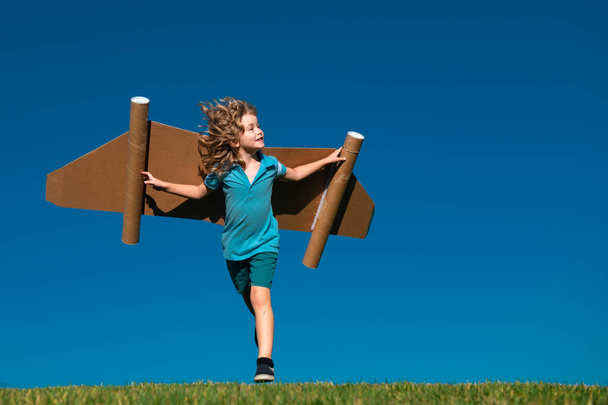 Το μικρό παιδί παίζει αστροναύτη ή πιλότο. Το παιδί τρέχει στο φόντο του γαλάζιου ουρανού. Παιδιά με χάρτινα φτερά ονειρεύονται. Τα παιδιά φαντάζονται όνειρα πτήσης. Αστείο παιδί με παιχνίδι τζετ πακέτο - Φωτογραφία, εικόνα
