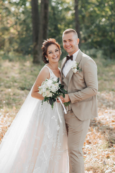 Wedding portrait. The bride and groom are standing in the forest hugging and looking at the camera, both holding a wedding bouquet. Long dress with train. Light suit of the groom. Beautiful makeup - Photo, Image