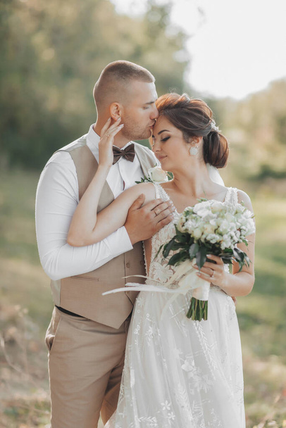 Wedding photo in nature. The bride and groom are standing in the forest, the groom hugs his beloved from behind and kisses her, she smiles sincerely. Portrait. Summer wedding - Photo, image