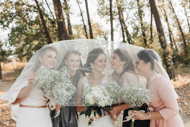 Wedding photo in nature. The bride and her friends are standing in the forest holding their bouquets, smiling, covered by the bride's veil. Wedding portrait. Emotions. Beautiful girls. - Fotoğraf, Görsel