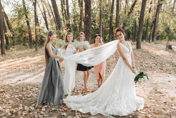 Wedding photo in nature. The bridesmaids stand behind the bride, holding their bouquets and her veil, the bride holds the bouquet and looks down. Wedding portrait. Emotions. Beautiful girls. - Valokuva, kuva