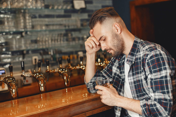 Man in a shirt holds a glass in his hands. Guy is sitting at the bar and holding his head. - Foto, imagen