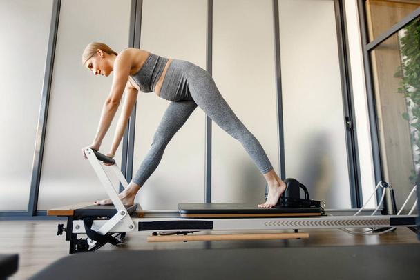 Pregnant woman doing exercises with a special equipment. Fit blonde woman working out in a gym. Woman wearing grey sportive leggins and top. - Foto, immagini