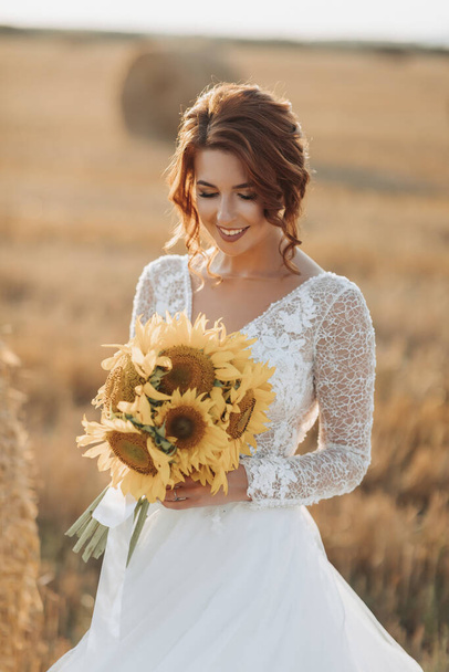 Wedding portrait. A red-haired bride in a white dress, holding a bouquet of sunflowers, on the background of a field. Beautiful curls. Sincere smile. Elegant dress. Yellow flowers - Photo, Image