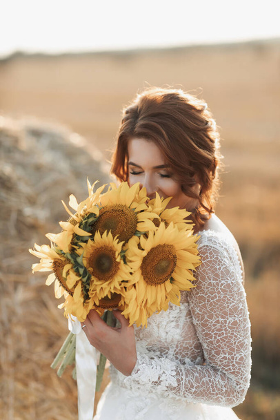 Wedding portrait. A red-haired bride in a white dress stands near a bale of hay with a bouquet of sunflowers and smells the flowers. Beautiful curls. Sincere smile. Elegant dress - Photo, Image