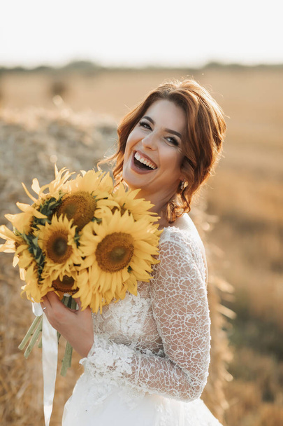 Wedding portrait. A red-haired bride in a white dress stands near a bale of hay with a bouquet of sunflowers and looking into the lens, smiling. Beautiful curls. Sincere smile. Elegant dress - Photo, Image