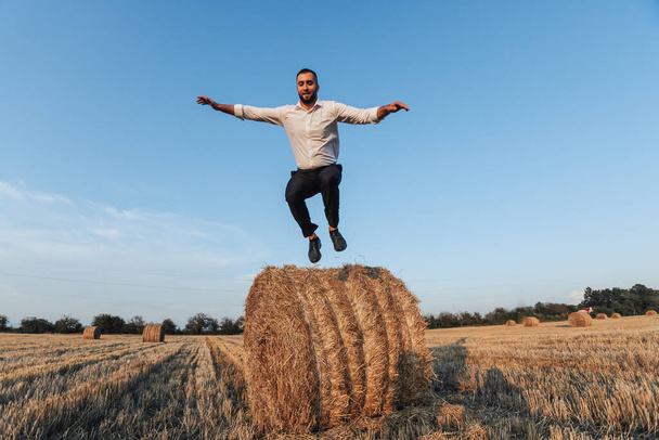 Wedding photo. Stylish groom in white shirt jumping down from hay bale and looking at camera. Bearded man. Style. Emotions - Valokuva, kuva