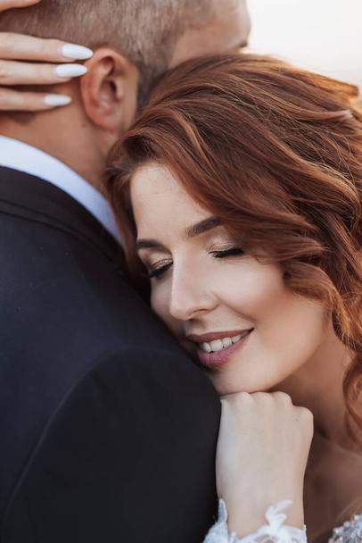 Wedding portrait. A red-haired bride in a white dress stands near a bale of hay with a bouquet of sunflowers and looking into the lens, smiling. Beautiful curls. Sincere smile. Elegant dress - Foto, Imagem