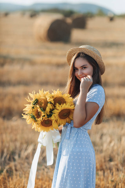 Portrait of a young girl. A girl in a blue dress and hat, holding a bouquet of sunflowers, against a background of hay bales. Long straight hair. Nice color. Summer - Zdjęcie, obraz