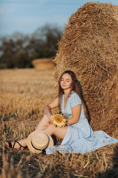Portrait of a young girl. A girl in a blue dress holds a sunflower flower against a background of hay bales. Long straight hair. Nice color. Summer - Foto, afbeelding