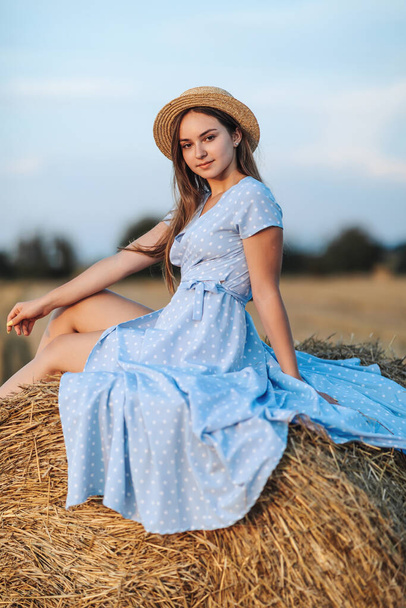 Portrait of a young girl. A girl in a blue dress sits on top of hay bales - high quality photo. Long straight hair. Nice color. Summer. Portrait - Foto, imagen