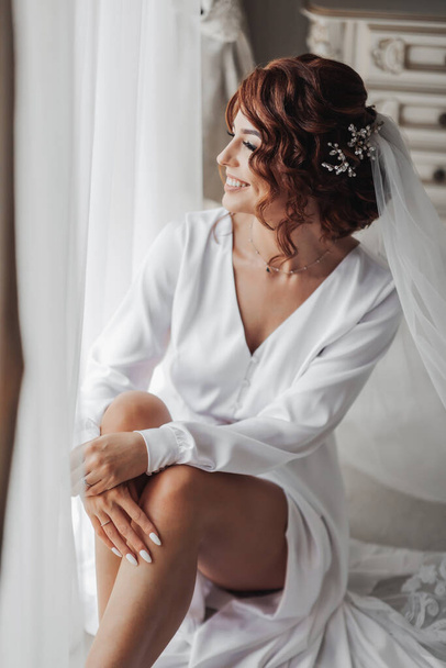 Fashion shot, beautiful woman in a dressing gown. Fashion, glamor concept. The morning of the bride, the bride in a dressing gown sits by the window posing for the photographer - 写真・画像
