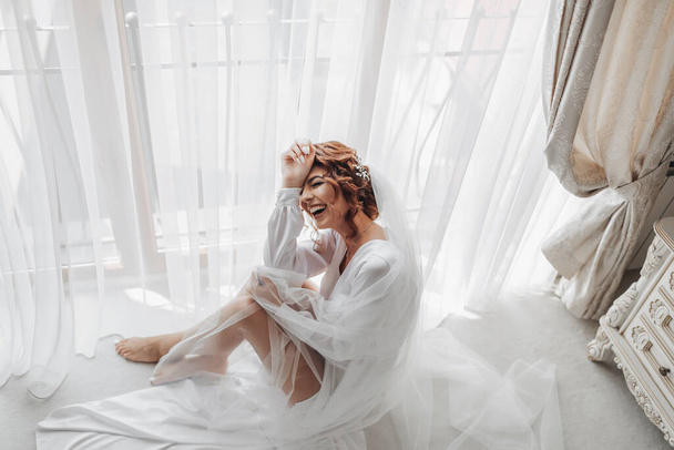 Fashion shot, beautiful woman in a dressing gown. Fashion, glamor concept. The morning of the bride, the bride in a dressing gown sits by the window posing for the photographer, smiling sincerely - 写真・画像