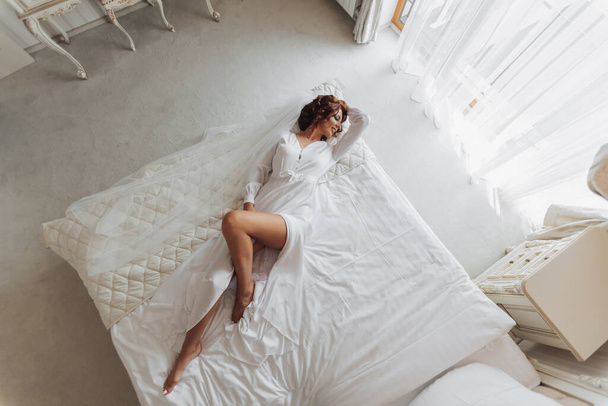 Belle jeune mariée en peignoir blanc couchée sur le lit dans la chambre d'hôtel. Derniers préparatifs du mariage. La mariée attend le marié. Bonjour, jeune mariée. Photo d'en haut - Photo, image
