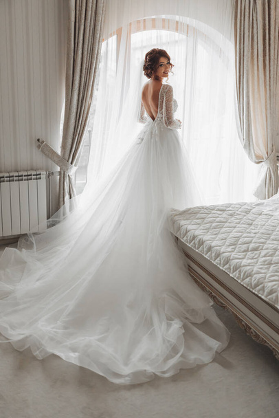 The bride in a beautiful dress is standing by the window with a long train. Wedding. wedding bouquet Morning of the bride. A happy bride is waiting for the groom. - Foto, Imagen