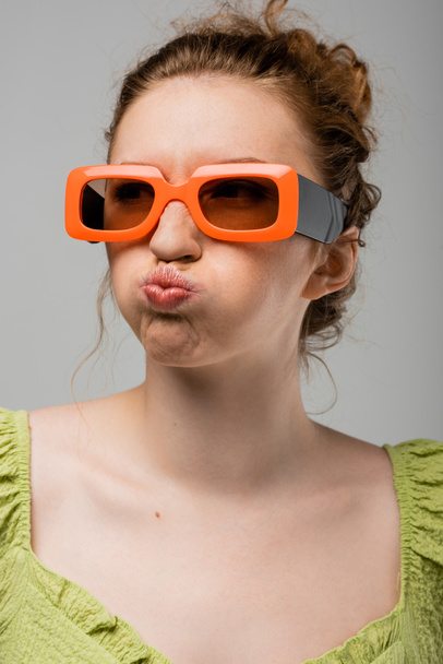 Retrato de mujer pelirroja joven en gafas de sol y blusa verde haciendo muecas y pucheros labios mientras está de pie aislado sobre fondo gris, concepto de protección solar de moda, modelo de moda  - Foto, Imagen