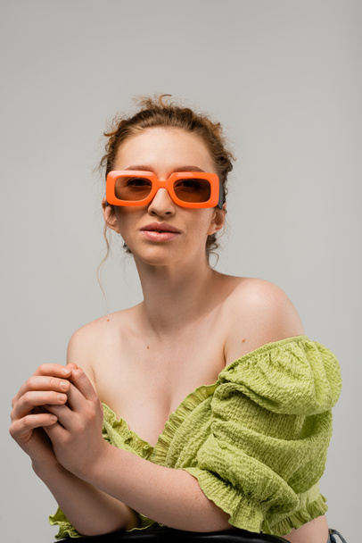 Portrait of stylish young red haired woman in sunglasses and green blouse with naked shoulders posing while standing isolated on grey background, trendy sun protection concept, fashion model  - Photo, Image