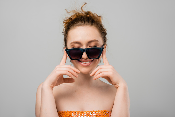 Fröhliche junge rothaarige Frau mit natürlichem Make-up in Sonnenbrille und orangefarbenem Top mit Pailletten, die Augen schließen, während sie isoliert auf grauem Hintergrund steht, trendiges Sonnenschutzkonzept, Modemodel  - Foto, Bild