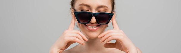 Retrato de mujer joven y sonriente con maquillaje natural y hombros desnudos tocando gafas de sol mientras está de pie aislado sobre fondo gris, concepto de protección solar de moda, modelo de moda, pancarta  - Foto, Imagen