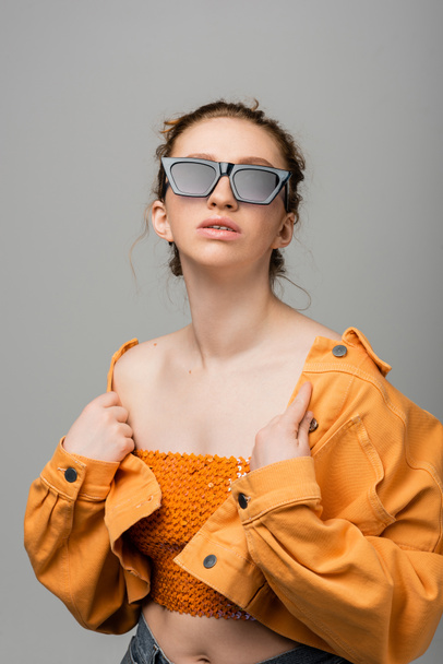 Fashionable red haired model in sunglasses and orange top with sequins touching denim jacket while standing and posing isolated on grey background, trendy sun protection concept, fashion model  - Fotó, kép
