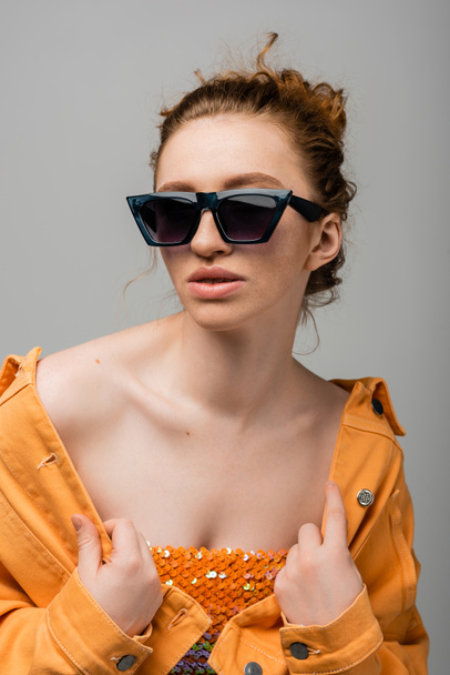 Young and redhead woman in sunglasses and top with orange sequins touching orange denim jacket and posing isolated on grey background, trendy sun protection concept, fashion model  - Photo, Image