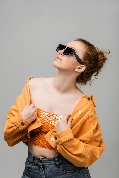 Stylish young redhead woman in sunglasses and top with sequins touching orange denim jacket and standing and posing isolated on grey background, trendy sun protection concept, fashion model  - Fotó, kép