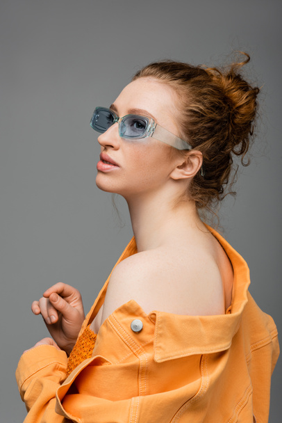 Fashionable young red haired woman in sunglasses and orange denim jacket with naked shoulder looking away and standing isolated on grey background, trendy sun protection concept, fashion model  - Photo, Image