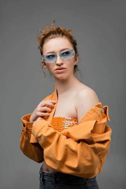 Stylish young red haired woman in sunglasses, top with sequins and orange denim jacket with naked shoulder standing isolated on grey background, trendy sun protection concept, fashion model  - Photo, Image