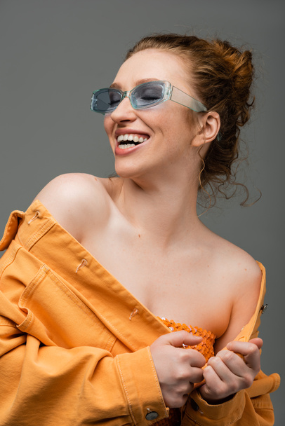 Wesoła i stylowa rudowłosa kobieta w niebieskich okularach przeciwsłonecznych i pomarańczowej kurtce jeansowej z nagimi ramionami śmiejąca się na szarym tle, modna koncepcja ochrony przeciwsłonecznej  - Zdjęcie, obraz