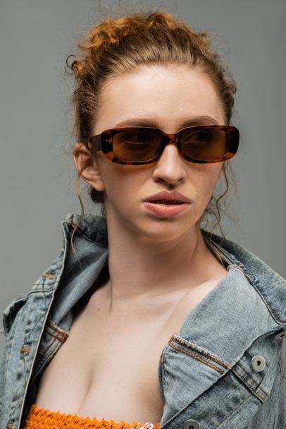 Confident young red haired woman with natural makeup wearing sunglasses and denim jacket and looking away isolated on grey background, trendy sun protection concept, fashion model  - Foto, Imagem