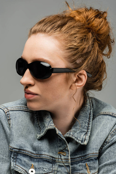 Portrait of stylish freckled and red haired woman in sunglasses and denim jacket posing and standing isolated on grey background, trendy sun protection concept, fashion model  - Photo, Image