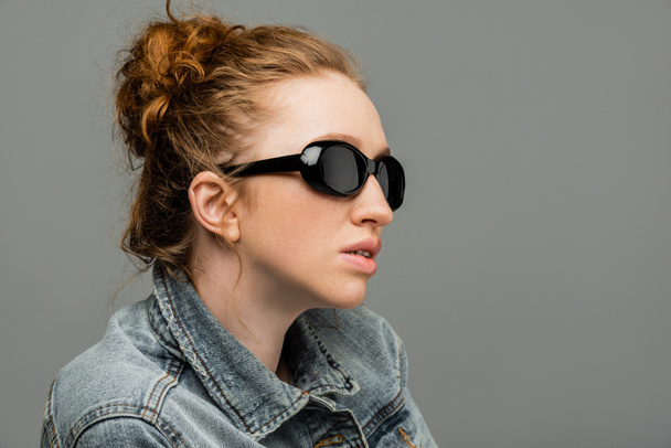 Fashionable young red haired woman with natural makeup wearing sunglasses and denim jacket while standing isolated on grey background, trendy sun protection concept, fashion model  - Photo, Image