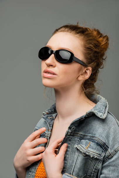 Κόκκινο μαλλιά και φακίδες νεαρή γυναίκα σε μοντέρνα γυαλιά ηλίου ποζάρουν σε τζιν σακάκι και κορυφή με πούλιες και στέκεται απομονωμένη σε γκρι φόντο, μοντέρνα έννοια αντηλιακή προστασία, μοντέλο μόδας  - Φωτογραφία, εικόνα