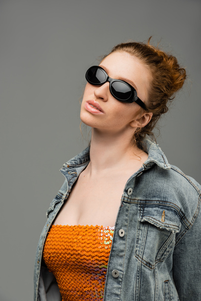 Confident and stylish young redhead woman in sunglasses, top with sequins and denim jacket standing and posing isolated on grey background, trendy sun protection concept, fashion model  - Fotó, kép