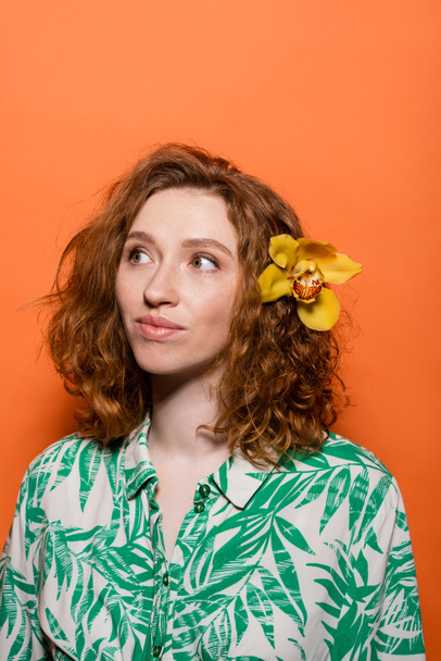 Junge rothaarige und sommersprossige Frau mit Orchideenblume im Haar trägt Bluse mit floralem Print und schaut weg auf orangefarbenem Hintergrund, Sommer-Freizeit- und Modekonzept, Jugendkultur - Foto, Bild
