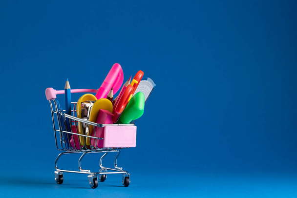 Close up of miniature shopping trolley with school materials and copy space on blue background. Back to school, shopping, school materials, learning, school and education concept. - Foto, Bild