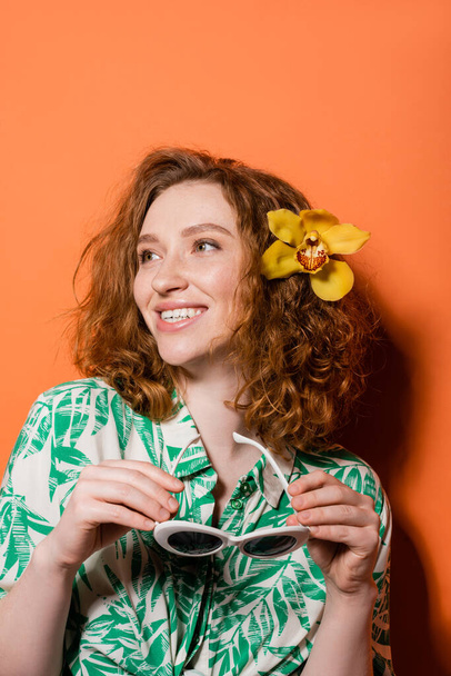 Uśmiechnięta młoda ruda kobieta z kwiatem orchidei we włosach nosząca bluzkę z kwiatowym nadrukiem i trzymająca okulary przeciwsłoneczne stojąc na pomarańczowym tle, letnia koncepcja casualu i mody, kultura młodzieżowa - Zdjęcie, obraz